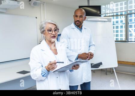 Leitende Ärztin erklärt Kollegen ein Gesundheitsprojekt, hält das Klemmbrett und zeigt mit einem Stift, steht neben einem männlichen Arzt in einem Betrüger Stockfoto