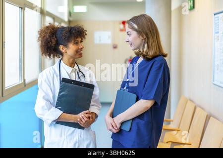 Ärztin und Krankenschwester führen ein freundliches Gespräch, während sie medizinische Unterlagen in einem geschäftigen Krankenhauskorridor durchsehen Stockfoto