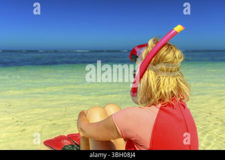 Blonde kaukasische Frau mit Schnorchelanzug, Maske und Flossen rosa und pfirsichfarbene, am Meer sitzend. Weiblich aussehendes tropisches Meer in Hangover B Stockfoto