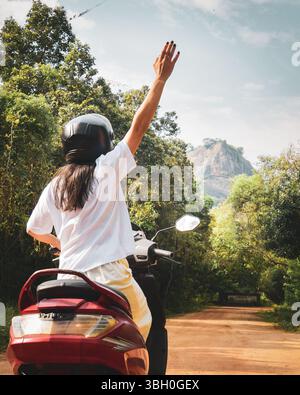 Zurück Ansicht Frau auf gemietetem Roller Fahrt winkend Besuchen Sie Pidurangala und Sigiriya Rock in Sri Lanka Abenteuer Road Trip durch tropischen Wald in sonnigem d Stockfoto