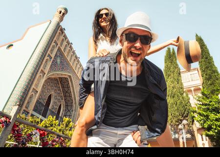 Fröhliche heterosexuelle Paare teilen sich Huckepack-Fahrt in der farbenfrohen Stadt, lachen und genießen helle Sommerurlaubsmomente. Konzept: Verbindung, Glück Stockfoto