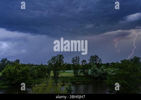 Blitze erleuchten einen düsteren Himmel über Bäumen und einem Fluss in der dunklen Nacht, Bogen, Niederbayern Stockfoto