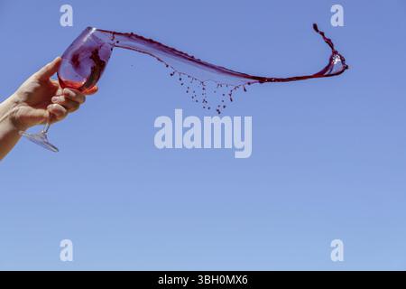 Die Hand hält ein Weinglas mit Rotwein, das vor einem klaren blauen Himmel spritzt Stockfoto