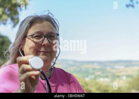 Reife Ärztin, die Stethoskop in der Natur hält und Gesundheit und Wohlbefinden fördert Stockfoto