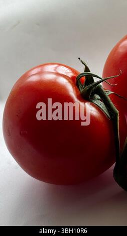 Nahaufnahme einer einzelnen Kirschtomate mit angenähtem Stiel, auf einer glatten weißen Oberfläche mit weichen Schatten. Stockfoto