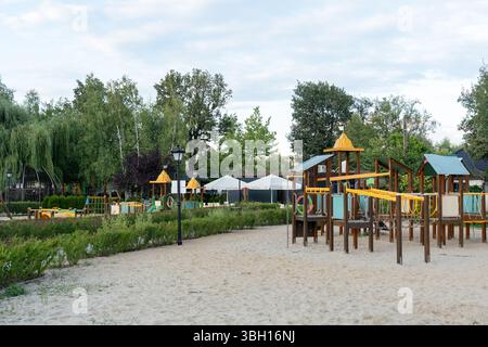 Moderner Spielplatz im Freien mit hölzernen Kletterstrukturen, Seilbrücken und Rutschen auf sandigem Untergrund im Park, umgeben von Bäumen. Das Konzept der Kinder Stockfoto