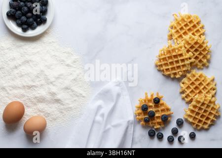 Blick von oben auf eine weiße Marmorplatte mit Zutaten für hausgemachte Waffeln. Neben Mehl und Eiern gibt es Heidelbeeren und fertige Waffeln Stockfoto