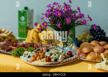 Ein lebhaftes Stillleben zeigt eine Käse-, Obst- und Snackplatte mit einer Saftbox, Blumen und Trauben, perfekt für Partys oder gesundes Essen Stockfoto