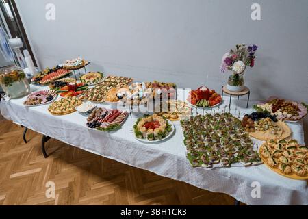 Eine lange weiße Tischdecke bietet ein umfangreiches Catering-Angebot mit farbenfrohen Vorspeisen, frischem Obst, Gemüse, Käse und Partydekorationen für Stockfoto