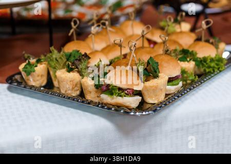 Ein Blick aus der Nähe auf köstliche Mini-Sandwiches und Vorspeisen, die kunstvoll auf einem eleganten silbernen Tablett angeordnet sind, perfekt für Catering, Bankette oder Cocktailrece Stockfoto