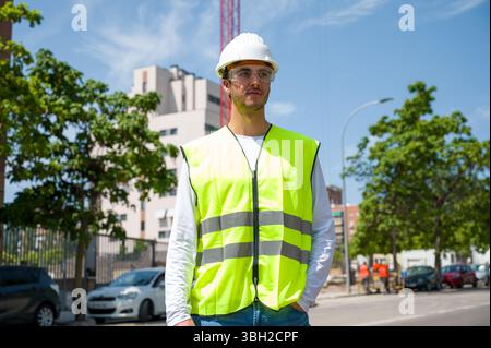 Professioneller junger Bauarbeiter mit Schutzhelm und reflektierender Weste auf einer Baustelle mit Baufortschritt im Hintergrund Stockfoto
