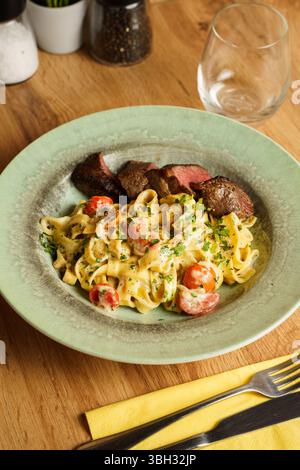 Ein Gourmetgericht mit cremiger Tagliatelle-Pasta, gemischt mit Kirschtomaten und frischen Kräutern, serviert mit saftigen Scheiben gegrilltem Steak. Das Gericht Stockfoto