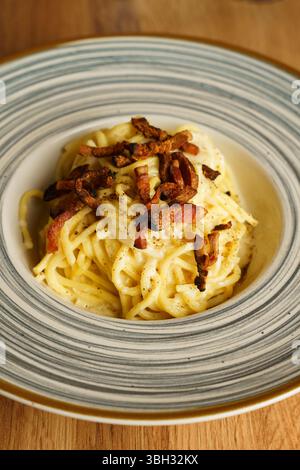 Eine wunderschön überzogene Portion Spaghetti Carbonara mit knusprigem Pancetta auf einer gestreiften Keramikplatte. Die cremige Soße und die goldenen Töne erzeugen einen Stockfoto