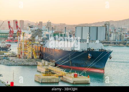 Großer LNG-Tanker (Flüssigerdgas), der an einem Industriehafen mit gelben Pipelines, Lagertanks und Kränen vor Anker liegt Stockfoto