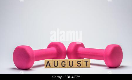 Holzblöcke mit der Schreibweise „August“ vor einer rosafarbenen Kurzhantel auf weißem Hintergrund Stockfoto