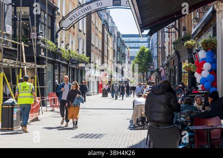 London, UK, 5. Mai 2025, Käufer und Touristen auf der Carnaby Street. Ein hell gekleideter Mann und eine Frau gehen an Bauarbeitern vorbei, die ein Haus renovieren Stockfoto