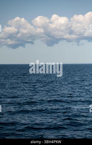 Ein kleines Segelboot segelt friedlich über das weitläufige blaue Wasser unter einem wolkenreichen Himmel. Stockfoto