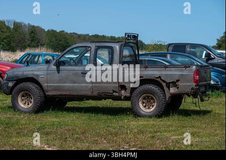 Rostiger Pickup-Truck sitzt auf grasbewachsenem Gelände mit verschiedenen Fahrzeugen unter klarem blauem Himmel. Stockfoto