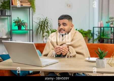 Kranker, kranker indischer Mann, der an einem Laptop arbeitet, der in Karo gewickelt ist, sitzt zitternd vor kaltem trinken heißem Kaffee im unbeheizten Heimbüro, ohne Schulden zu heizen. Ungesunder Araber versuchen, sich aufzuwärmen Stockfoto