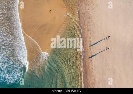 Blick von oben auf Paare, die am Sandstrand entlang spazieren, der von den Wellen des Atlantiks in Portugal durchzogen ist und lange Schatten bei niedrigem Sonnenaufgang wirft. Aufnahme während der Th Stockfoto