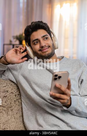 Junger indischer Mann auf dem Sofa, der kabellose Kopfhörer trägt Musik auf dem Smartphone hört wählt den Lieblingstitel entspannt und tanzt. Arabian Guy genießt Rhythmus, der sich mit Energie und fröhlichem Ausdruck bewegt. Stockfoto