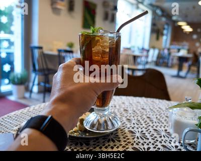 Die Person hält ein Glas Tee mit einem Strohhalm drin. Die Szene spielt in einem Restaurant mit Tischen und Stühlen Stockfoto