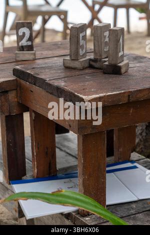 Holztisch mit drei Holzblöcken darauf, jeder mit einer Nummer. Der Tisch ist am Strand und es sind Stühle drumherum Stockfoto