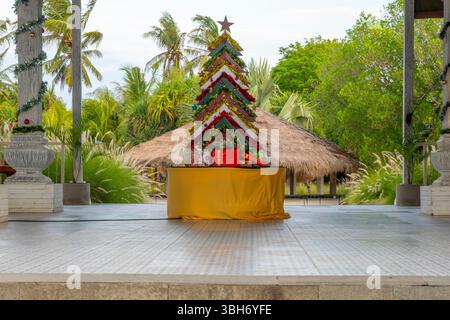 Weihnachtsbaum aus Holzstäbchen steht auf einer Bühne. Der Baum ist mit Lichtern und Geschenken dekoriert. Die Bühne ist von Palmen und einer Hütte umgeben. T Stockfoto