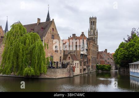 Der berühmte mittelalterliche Glockenturm von Brügge erhebt sich über dem malerischen Wasser des Kanals, mit traditionellen Backsteinbauten und Bäumen im Vordergrund. Stockfoto