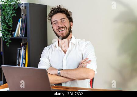 Ein fröhlicher junger Berufs- oder Unternehmer mit Bart lächelt selbstbewusst in die Kamera und sitzt mit überkreuzten Armen an seinem Schreibtisch mit einem Laptop in einem Stockfoto