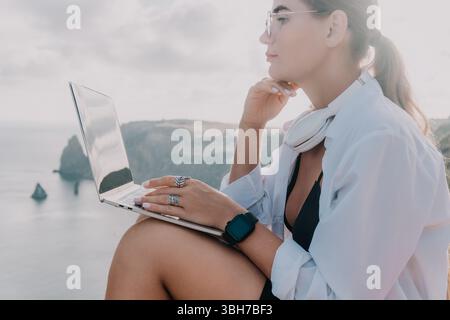 Woman Laptop Beach Working - Eine Frau sitzt auf einer Klippe mit Blick auf das Meer und arbeitet an ihrem Laptop. Stockfoto