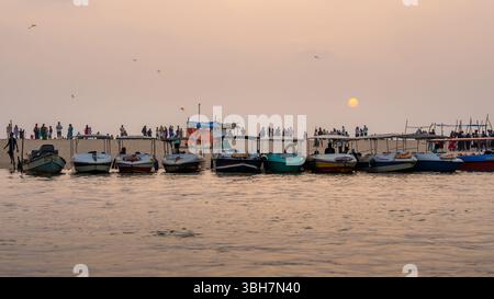 Boote ankerten bei Sonnenuntergang an einem beliebten Strand in Kerala, Indien, mit Menschenmassen und fliegenden Drachen im Hintergrund Stockfoto