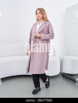Frau in einem langen, mauvenfarbenen Mantel geht selbstbewusst drinnen, trägt schwarze Hosen und dunkle Schuhe. Eine weiße Couch und ein hoher Spiegel sind im Raum, was einen schafft Stockfoto