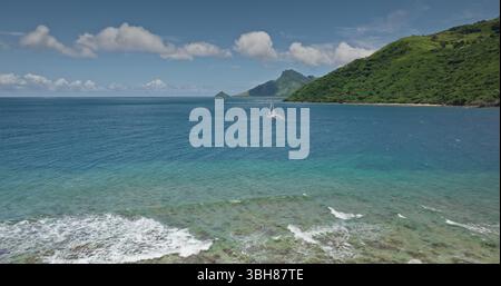 Türkisfarbene Wellen des tropischen Ozeans prallen gegen das pulsierende Korallenriff, Katamaran gleiten anmutig in die Nähe üppiger grüner Inseln am sonnigen Sommertag, malerisches Paradies. Fantastischer Naturreisehintergrund Stockfoto