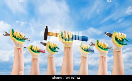 Die Hände des Mannes Arbeiter halten verschiedene Werkzeuge Hammer, Schraubendreher, Zange in Handschuhen vor blauem Himmel mit Wolken. Arbeitstag-Konzept. Stockfoto