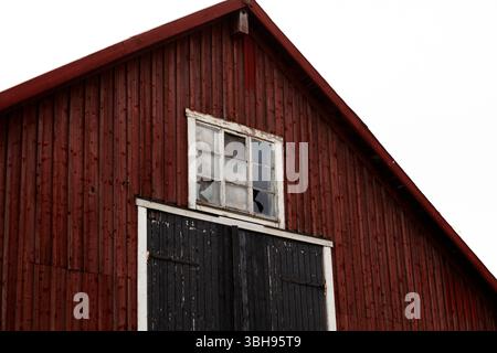 Aus nächster Nähe sehen Sie eine traditionelle skandinavische rote Holzscheune mit vertikalem Abstellgleis, einer schwarzen Holztür und einem kleinen weiß gerahmten Fenster unter dem Dach Stockfoto