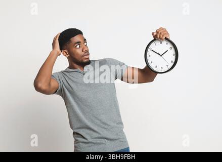 Schockierter schwarzer Mann, der eine Wanduhr auf weißem Hintergrund hält Stockfoto