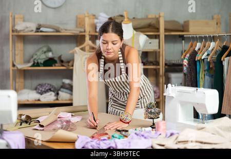 Die Schneiderin zeichnet in der Nähwerkstatt anhand von Mustern eine Skizze der zukünftigen Kleidung auf Papier Stockfoto