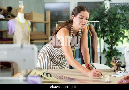 Die Schneiderin zeichnet in der Nähwerkstatt anhand von Mustern eine Skizze der zukünftigen Kleidung auf Papier Stockfoto