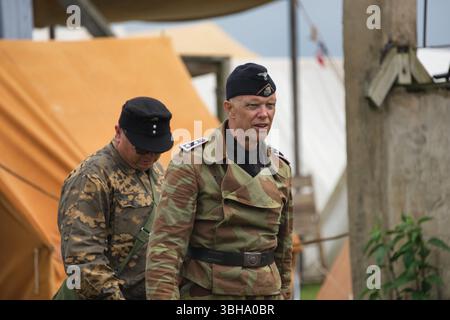 Zwei Männer in deutschen Militäruniformen aus dem Zweiten Weltkrieg stehen draußen neben einem braunen Zelt und einem Betonpfosten. Ein Mann trägt eine Tarnjacke. Stockfoto
