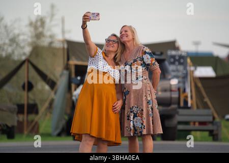 Zwei Frauen in Kleidern machen ein Selfie. Das eine trägt ein senfgelbes Kleid, das andere ein Kleid mit Blumenmuster. Sie stehen draußen vor verschwommenem Militär Stockfoto