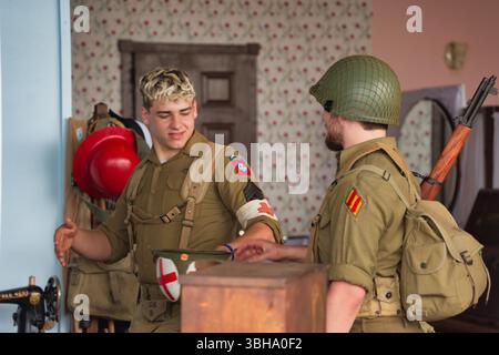 Zwei Männer in Uniformen der US-Armee interagieren in der Nähe einer Holzkiste. Einer trägt ein medizinisches Armband. Ein Gewehr und andere Punkte sind im Hintergrund sichtbar Stockfoto