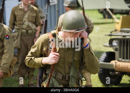 Ein Soldat in einer Uniform der US-Armee aus dem Zweiten Weltkrieg benutzt ein Feldtelefon. Er trägt ein Gewehr und andere Ausrüstung. Zwei weitere Soldaten sind im Hintern verschwimmen Stockfoto