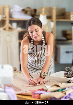 Die Schneiderin zeichnet in der Nähwerkstatt anhand von Mustern eine Skizze der zukünftigen Kleidung auf Papier Stockfoto