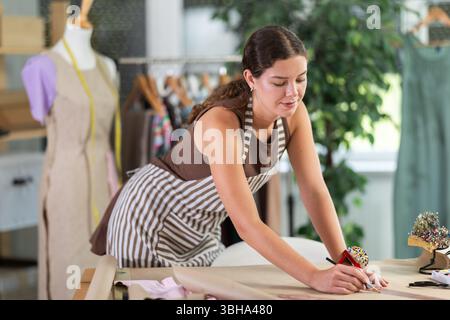 Die Schneiderin zeichnet in der Nähwerkstatt anhand von Mustern eine Skizze der zukünftigen Kleidung auf Papier Stockfoto