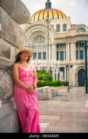 Glückliche Frau mit rosa Kleid und Strohhut, die sich an eine Steinmauer in der Nähe des Palacio de Bellas Artes in Mexiko-Stadt lehnt, mit dem legendären goldenen Do Stockfoto