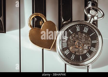 London, Großbritannien - 19. April 2025 - Ansicht von oben auf Eine Retro-Taschenuhr oder eine antike Taschenuhr mit goldenem Herz geformtem Liebesvorhängeschloss auf Klaviertasten Hintergrund in Stockfoto