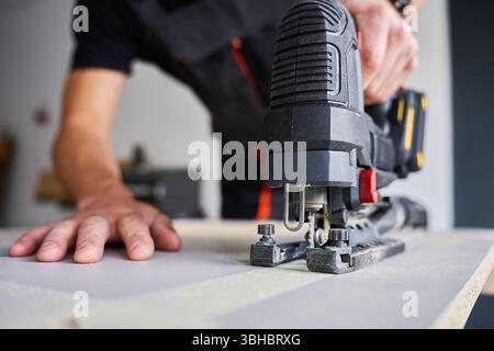 Arbeiter in Arbeitskleidung, der mit einer kabellosen Handstichsäge Holzbretter in der Garage schneidet. Geschulter Schreiner, der in der Werkstatt arbeitet. Ein Mann, der Heimwerker macht Stockfoto