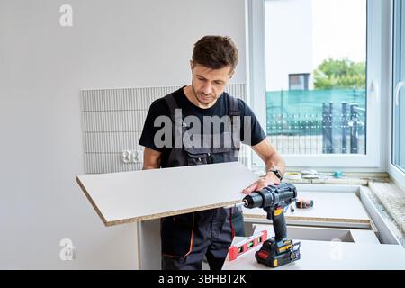 Mann in Uniform misst Schrankpaneel bei der Installation von Küchenmöbeln. Renovierungsarbeiten in der neuen Wohnung Stockfoto