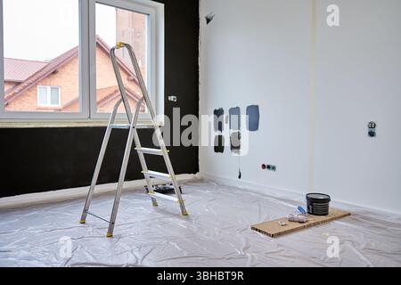 Der Raum wird gerade renoviert, mit Farbmustern an der weißen Wand, Leiter und Malwerkzeugen auf dem Boden mit Schutzfolie Stockfoto